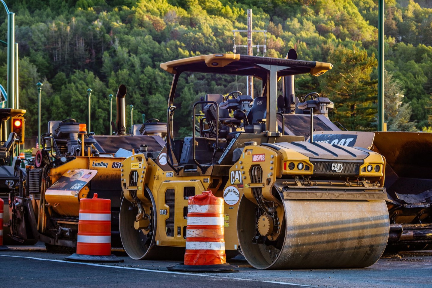 Yellow road rollers and paving equipment parked beside a highway, with a forested hillside behind.
