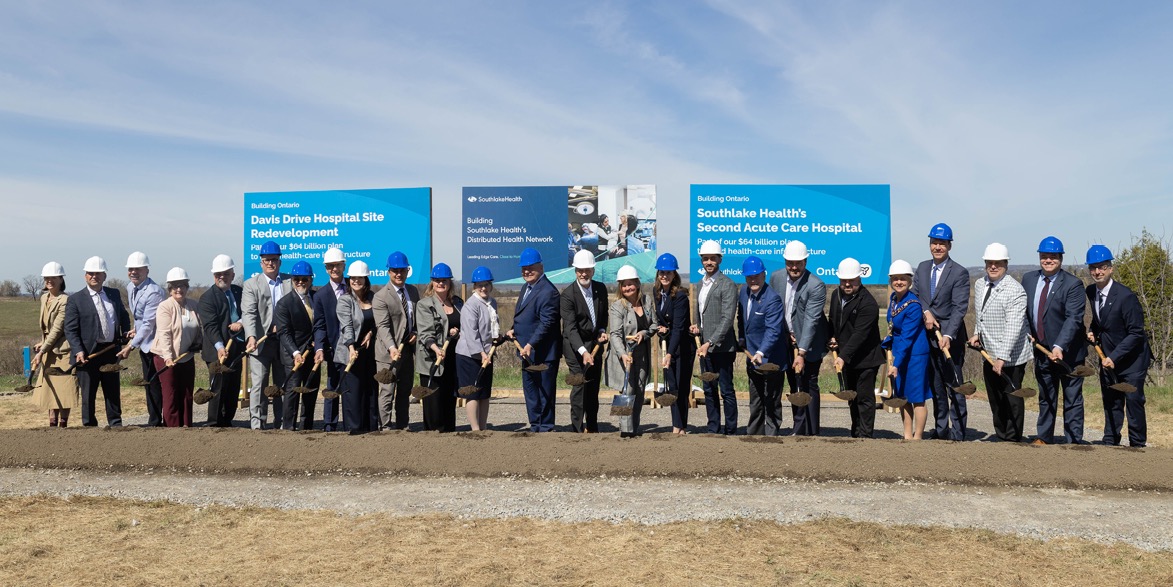 Officials in hard hats with ceremonial shovels at the East Gwillimbury site, in front of Southlake Health and Building Ontario signs for the second acute care hospital and the Davis Drive redevelopment.