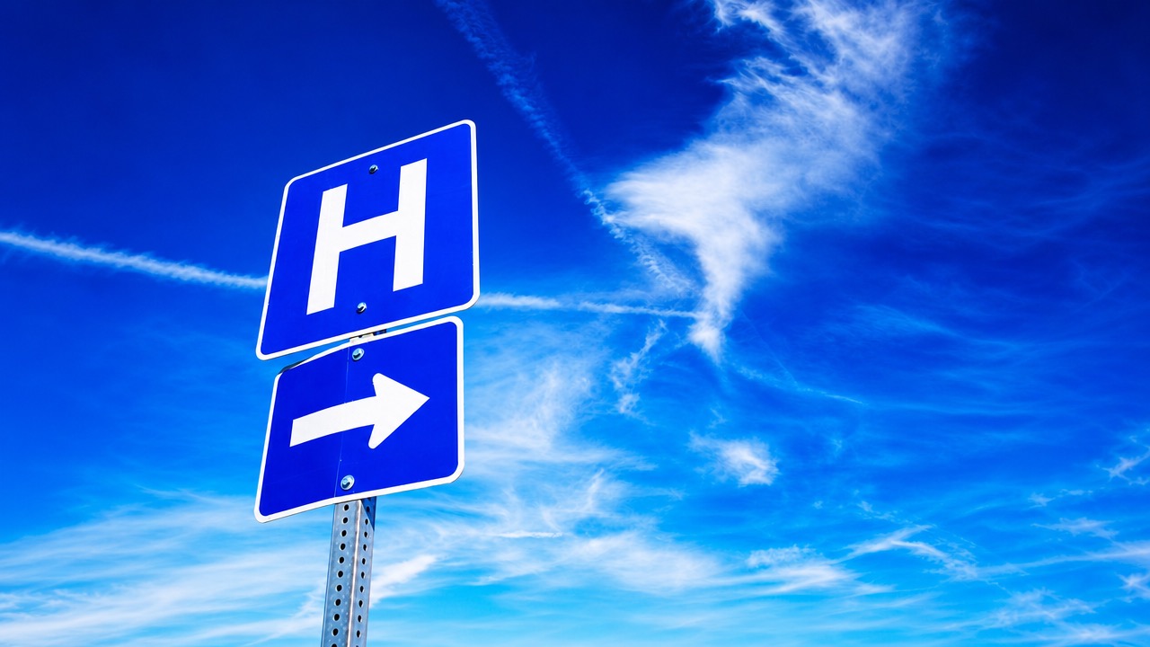 A blue hospital wayfinding sign with a white H and a directional arrow, against a bright blue sky.
