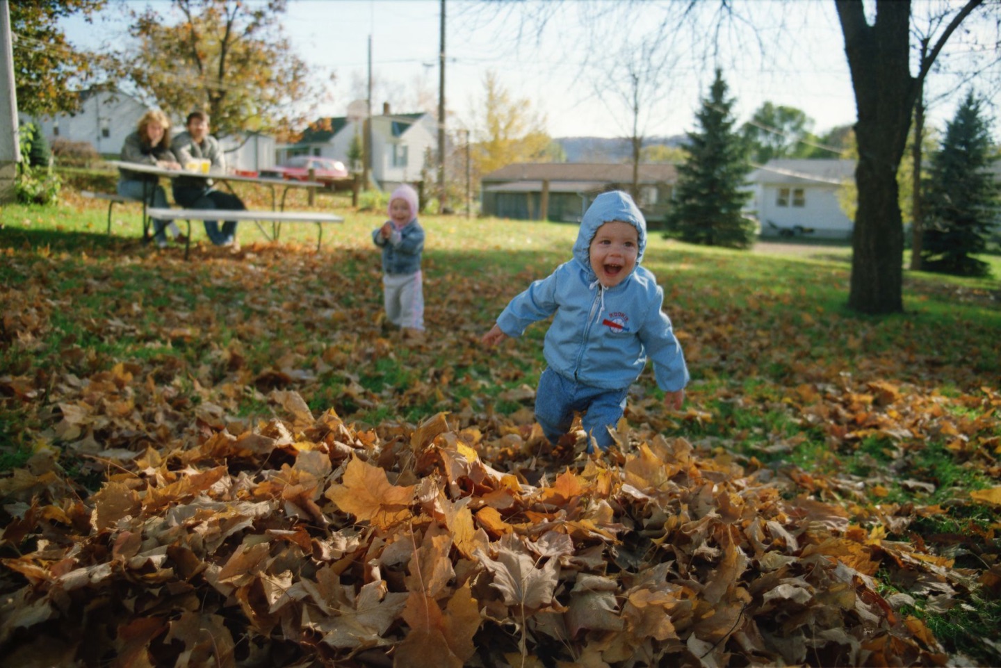 A young child in a blue jacket laughs while running through a pile of autumn leaves in a suburban park, with houses and a picnic bench behind.