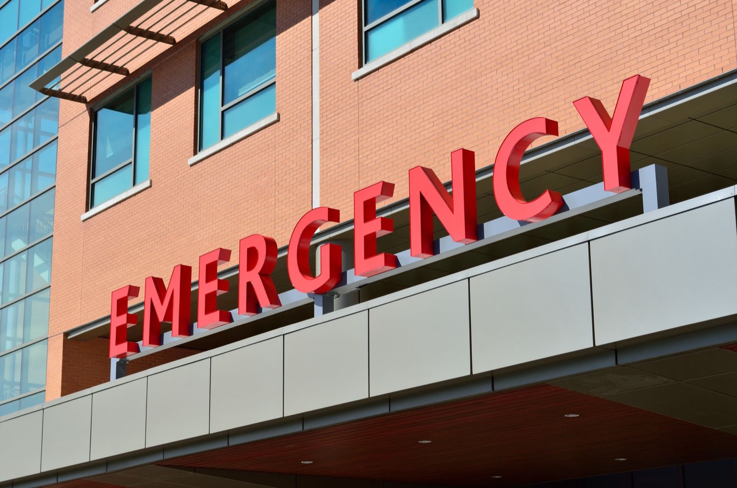 The red EMERGENCY sign on the exterior of a modern brick-and-glass hospital.